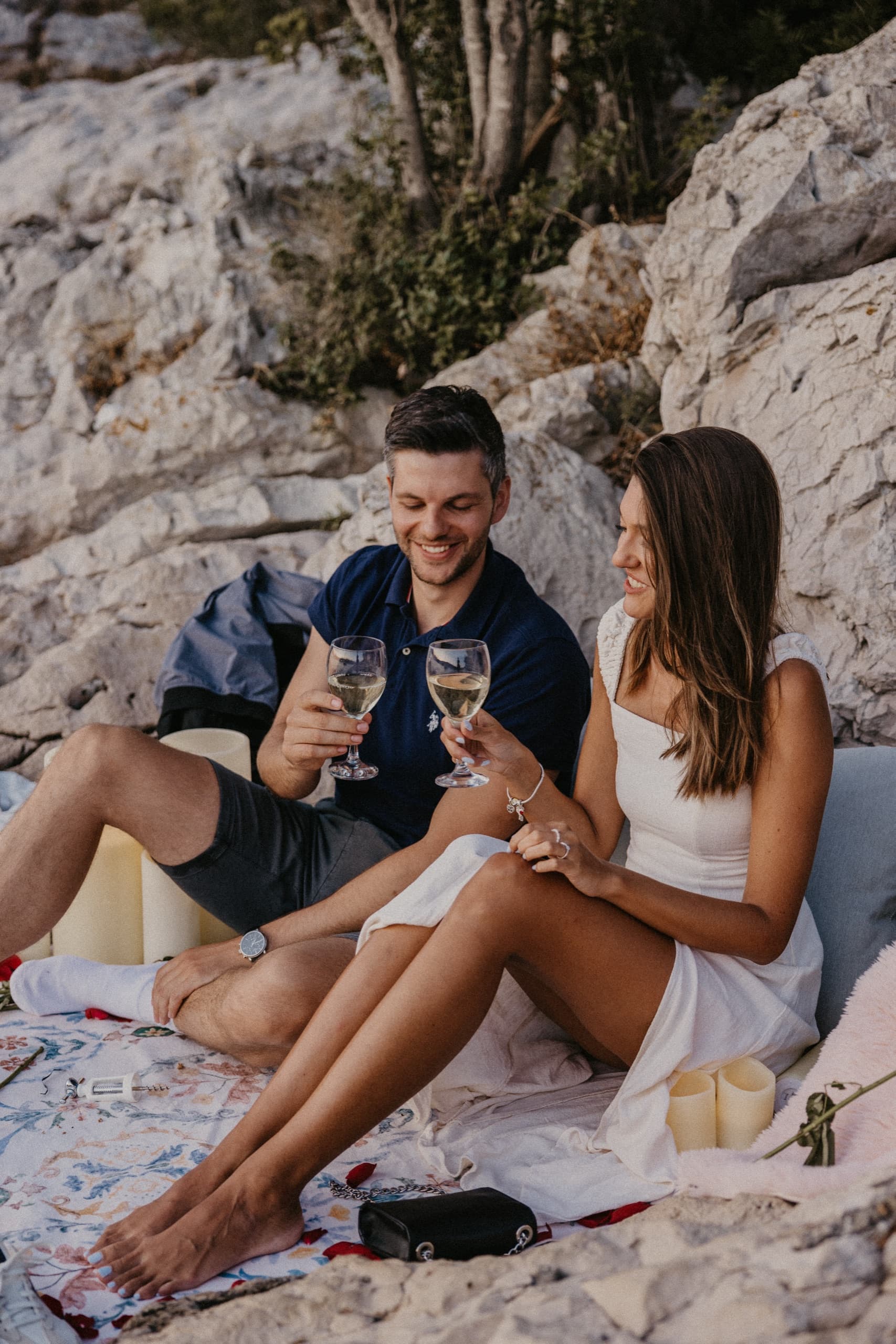 A couple laughing over glasses of wine on a rug by the sea in golden light