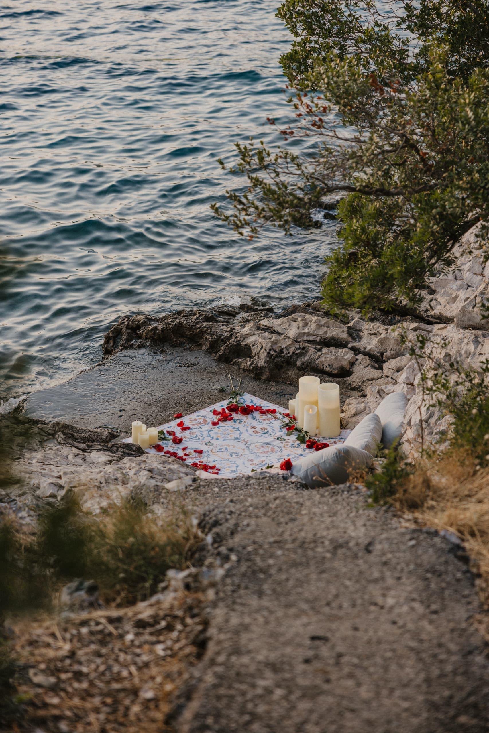 A rug scattered with rose petals and candles on the rocky shore at golden hour